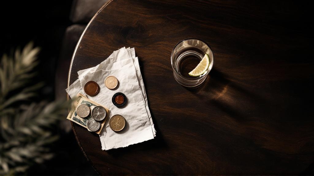Overhead view of a dark wooden bar table with an empty glass ring, crumpled receipt, cards, and loose coins symbolizing the hidden cost of a night out.
